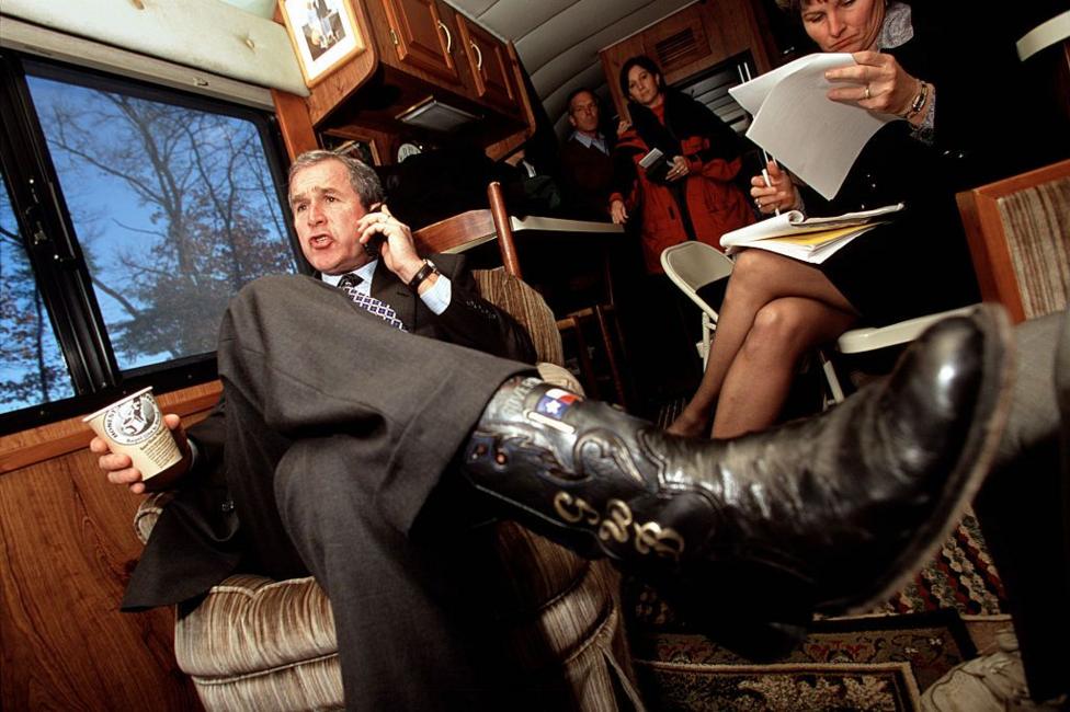 Former US president George W Bush seated inside a campaign-style bus, wearing a dark suit and patterned tie while talking on a mobile phone and holding a paper coffee cup. One leg is stretched forward, showing a black cowboy boot decorated with a Texas flag motif. Other people are visible in the background reviewing documents.
