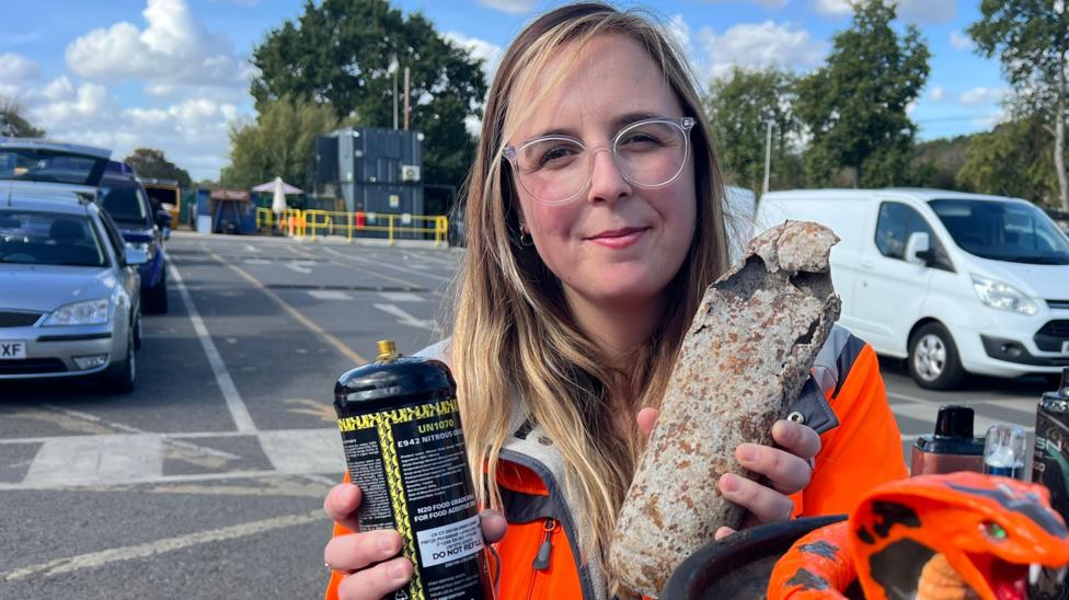 A blonde-haired woman wearing clear-framed glasses faces the camera. She holds a canister and an exploded shell, while wearing an orange high-visibility jacket. She is in a car park setting, with blue sky and white clouds above.