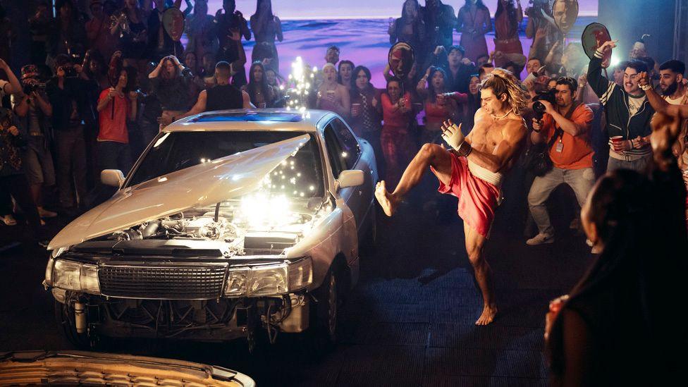 Noah Centineo's character launches a flurry of kicks at a silver saloon car as a large crowd watches and cheers him on. The bonnet of the car is flying off in a shower of white sparks.