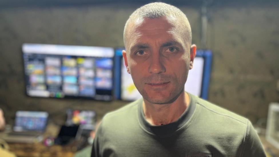 Man in Ukrainian military t-shirt poses in front of a set of computer desks with several screens. 