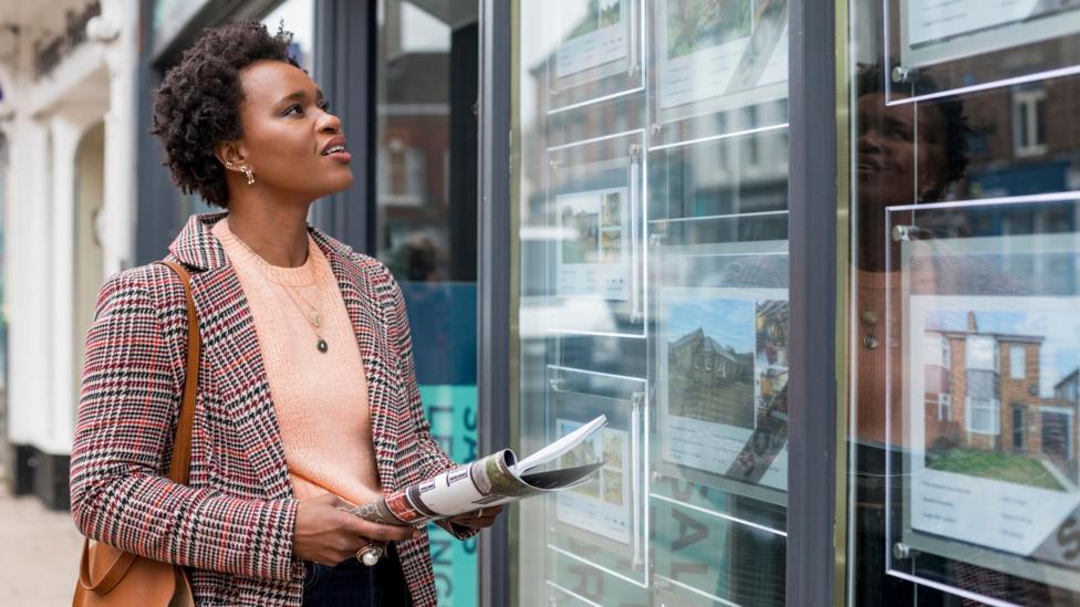 A stock image of a lady perusing an estate agent's available properties in the window. She carries a newspaper, a handbag, and a blazer.