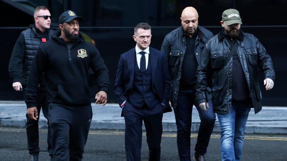 Tommy Robinson qo'llari cho'ntagida, xavfsizlik xodimlari bilan o'ralgan holda yurmoqda