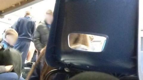 Этот снимок, сделанный из-под парты, показывает детей, собравшихся в классе. На переднем плане стул, закрывающий часть кадра. Лица детей размыты.
