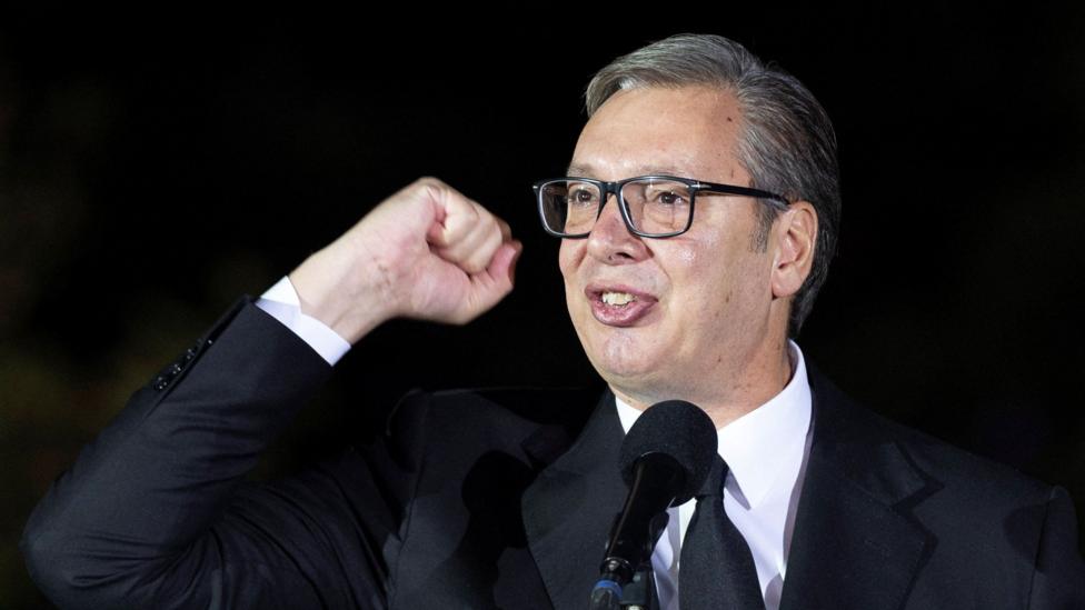 A head shot of Aleksandar Vucic looking off-camera giving a speech while waving his fist in the air. He is wearing a black suit and tie with a white shirt.