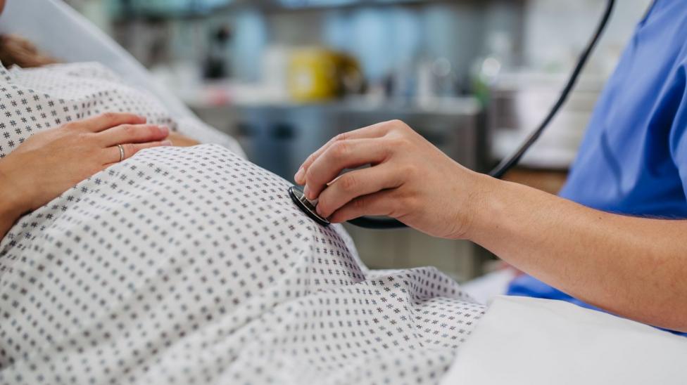 Close up of a pregnant belly with a medic in scrubs holding a stethoscope against the stomach.