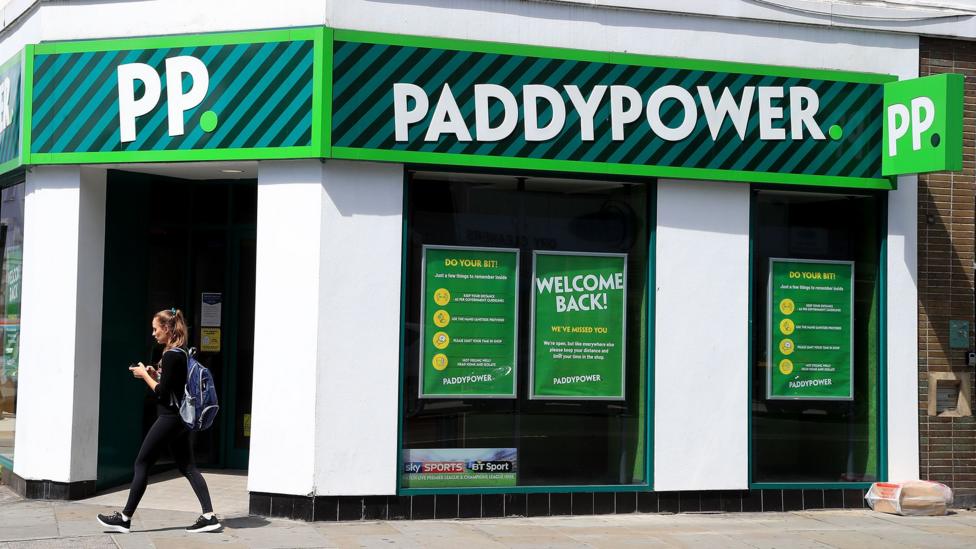 Логотип Paddy Power виден на вывеске филиала в Лондоне в 2020 году; в солнечный день по улице перед ним идёт человек, глядя в телефон.