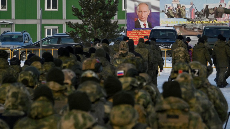 The backs of a crowd of soldiers wearing military camos with the Russian flag. They are facing a poster of Vladimir Putin