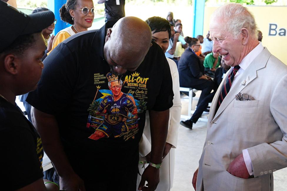 King Charles meets 12-year-old Sanai Miller and boxing coach Jermaine Gibson during a visit to The Bahamas Boxing Academy in Nassau. Gibson is wearing a black t-shirt with a cartoon version of Charles as a boxer on it.