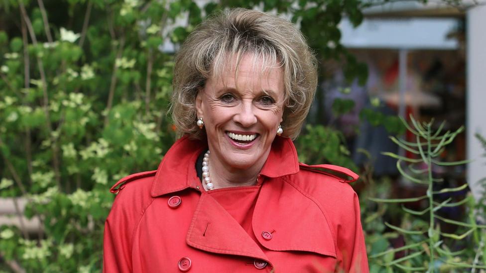 Head shot of Dame Esther Rantzen looking at the camera smiling. She's wearing a red coat, with pearl earrings and a matching pearl necklace, with green plants in the background.