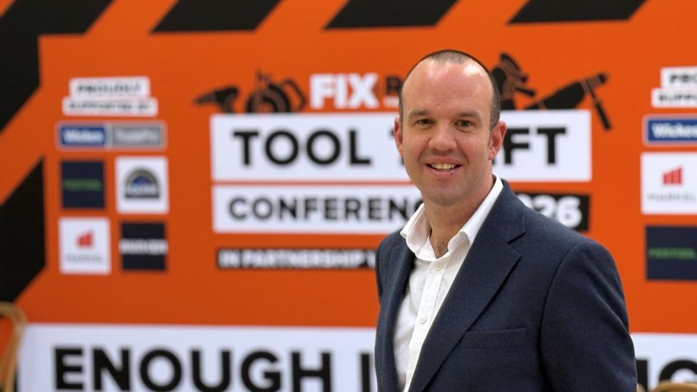 Gareth Powley kameraga yon tomondan qaragan va tişlari körinadigan tabassum bilan kulmoqda. U oçiq yoqali oq köylak va töq kök kostyum kiygan. Orqasida Fix Radio Tool Theft 2026 konferensiyasi belgisi va «Enough Is [Enough]» şiorining yarmi körinib turibdi.