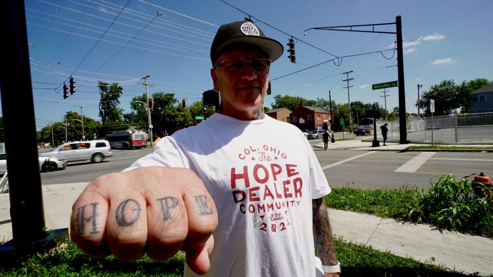 Scott Sanders, 65, wears a baseball cap and T-shirt that reads The Hope Dealer Community. He points his fist at the camera and on his knuckles reads Hope. 