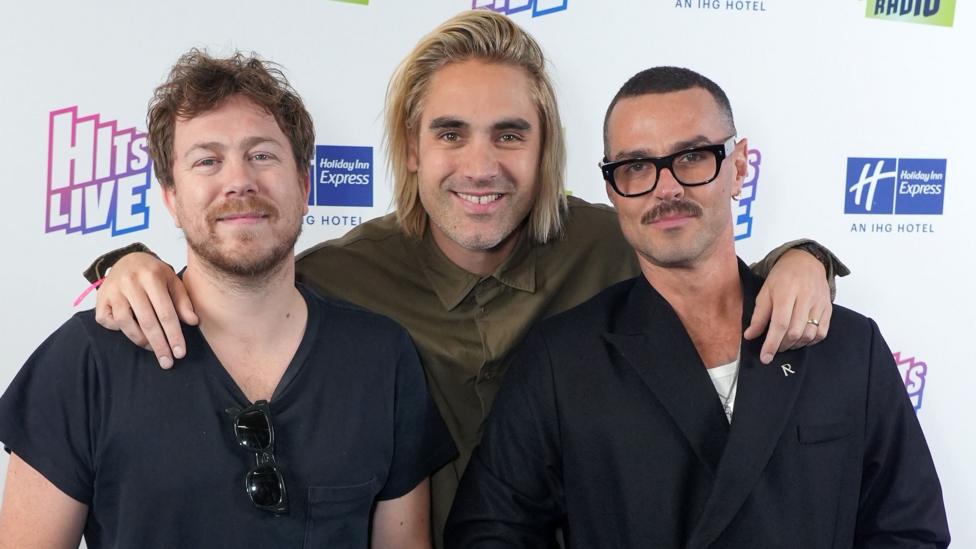 Bourne standing and posing with Charlie Simpson and Matt Willis in 2023; Simpson has his arms around the other two's shoulders