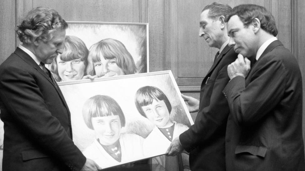A black and white photograph showing three men in suits looking at two paintings of some of the children who died in the Aberfan disaster