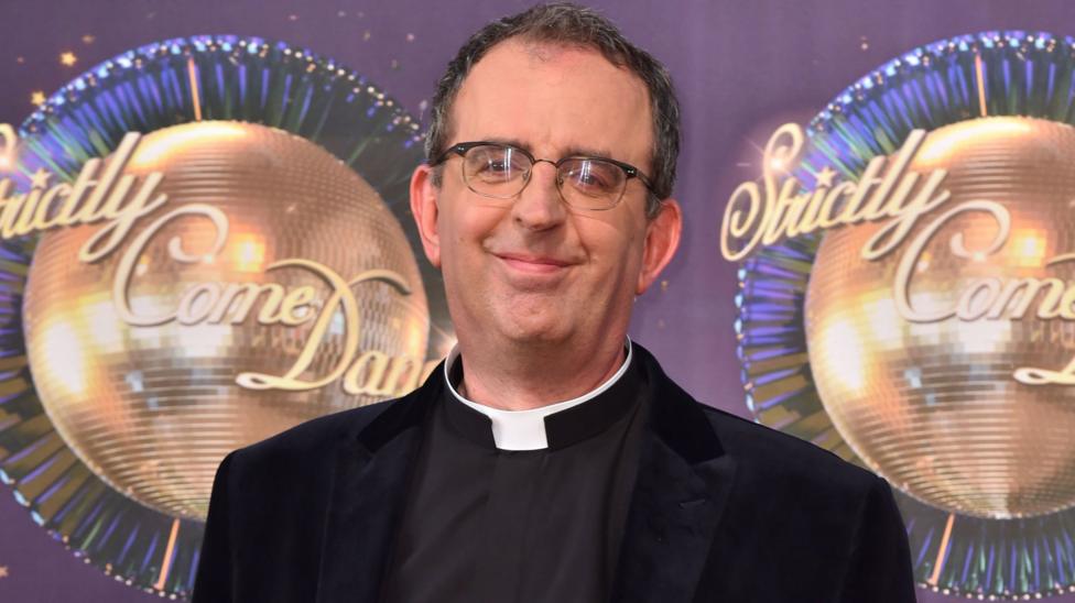 A head and shoulders image of Reverend Richard Coles who is wearing spectacles and a black suit with a clerical collar. He is standing in front of a backdrop with the Strictly Come Dancing logo and glitterballs.