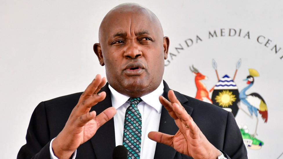 Uganda's Health Minister Chris Baryomunsi, in black suit and white shirt, gestures as he speaks. There is a black microphone in front of him and a banner with words Uganda Media Centre and a Ugandan coat of arms behind him.