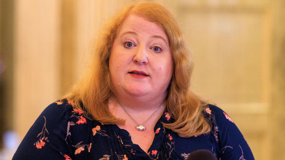 A woman with light hair, she has a navy top with pink flowers on it