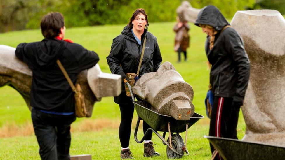 Miranda in a raincoat pushing a wheelbarrow with a stone head across the grass, in between two other celebrities and parts of statues
