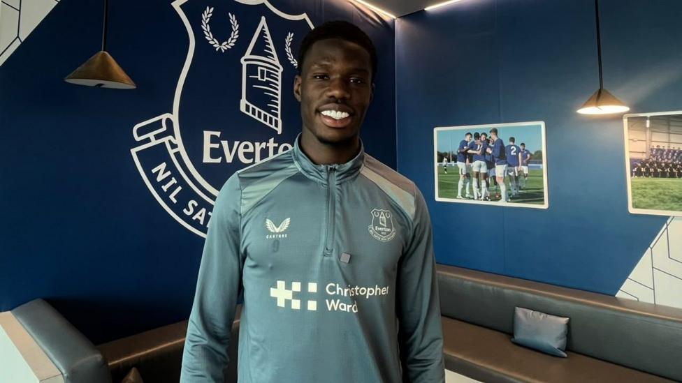 Thierno Barry stands up and smiles at the camera at Everton's training ground. He wears a light blue Everton sports top and has closely cut black hair. Behind him is a blue wall with the Everton logo, a grey sofa seat and pictures of players on the wall